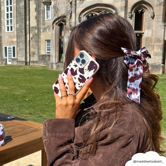 Woman with long hair and cow-print bow holding a Rodeo Cowgirl cow-print phone case outside a stone building.