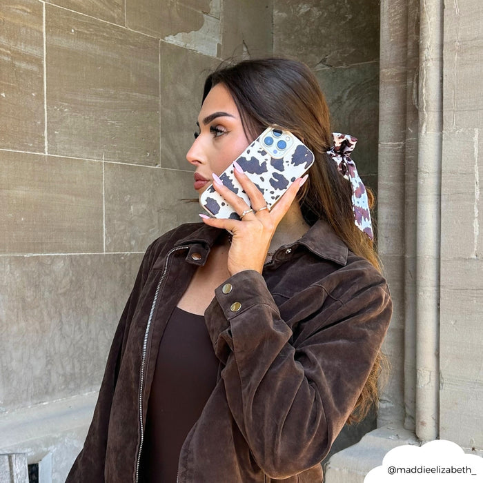 Woman with long hair in a printed hair tie holds a Rodeo Cowgirl phone case featuring a cow print, wearing a brown jacket.