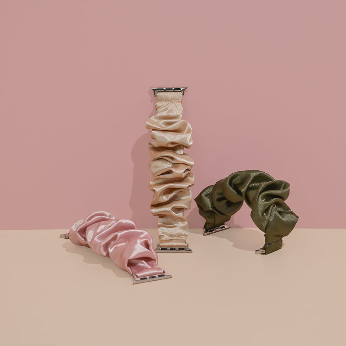 Three scrunchie-style watch bands from Coconut Lane are displayed on a two-toned background with a pink upper half and beige lower half. The Scrunchie Apple Watch Straps come in pink, beige, and olive green. The caramel latte scrunchie-inspired beige band stands upright while the others lay flat.
