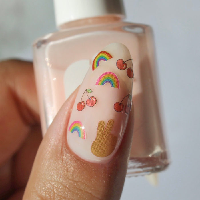 Serene Dreamin' rainbow, cherry and peace sign decals on a sheer nude nail, holding the bottle