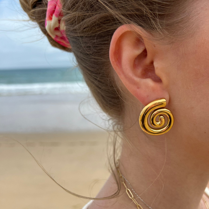 Close-up of Coconut Lane Shell Soirée earrings with a shell design on a womans ear, beach and ocean softly blurred behind.