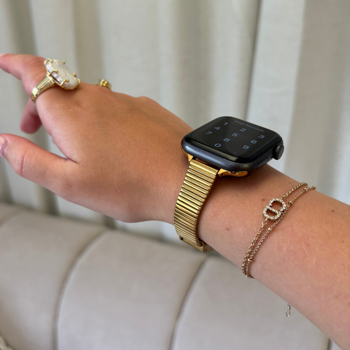 Gold SLIM Fine Line Apple Watch strap on wrist with gold rings and a delicate bracelet, hand resting near a beige couch.