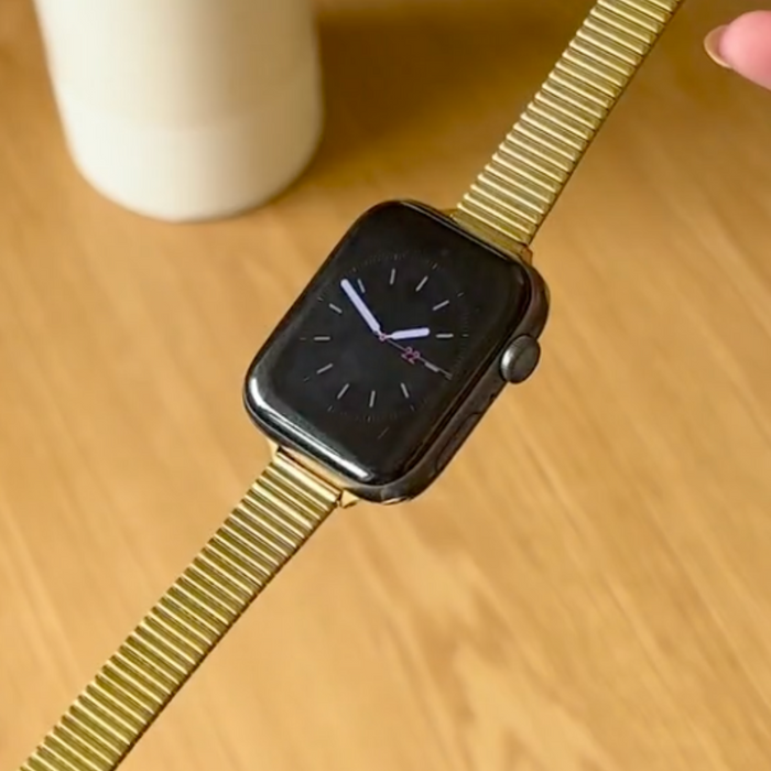 Black smartwatch with a gold fine line strap held above a wooden surface next to a cup.