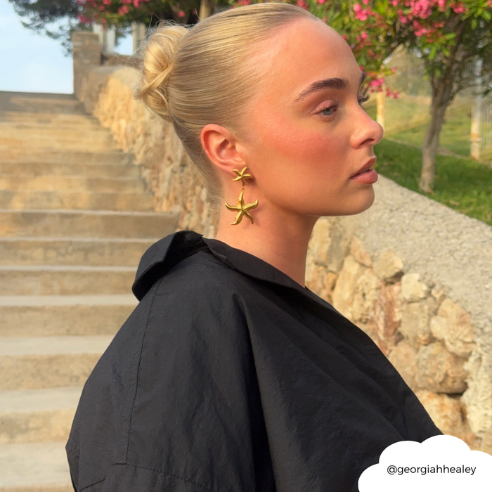 Woman with blonde hair in a bun wears gold starfish earrings and a black top outdoors by stone steps.