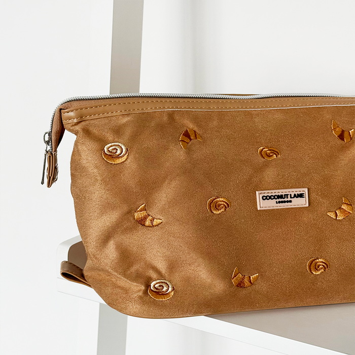 Tan suedette expandable makeup bag with croissant pastry motifs, shown on a white shelf.