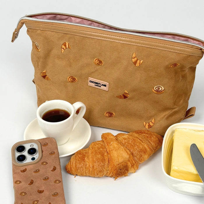 Beige suedette expandable makeup bag with croissant pattern, next to a croissant, coffee cup, and phone.