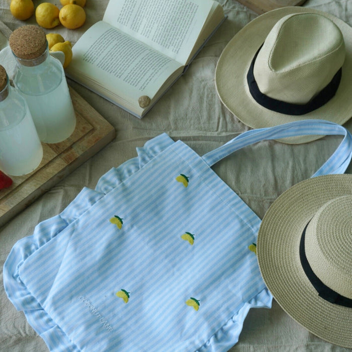Yellow Living La Vida Lemon tote bag by Coconut Lane with two hats, a book, and water bottles on a beige blanket.