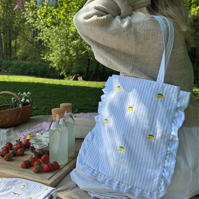 Woman holding yellow Living La Vida Lemon Coconut Lane tote bag at a picnic with strawberries and snacks on a tray.