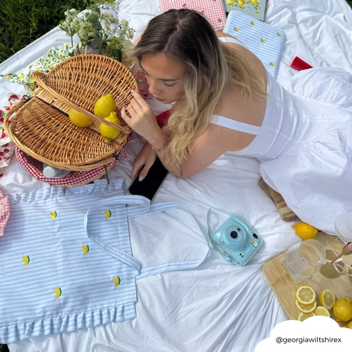 Woman in a white dress sits on grass beside lemons, a camera, and a yellow patterned Coconut Lane tote bag.