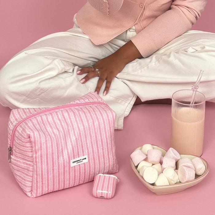 Woman sitting cross-legged with pink accessories, holding a striped pink Coconut Lane Airpods case and a pink drink.