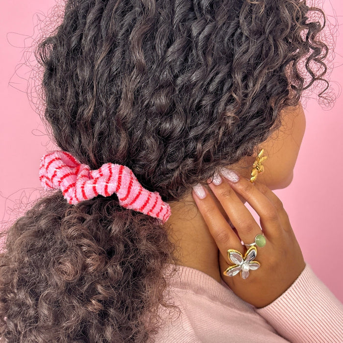 Woman with curly hair wearing a Cherry Stripes patterned towel scrunchie and gold jewelry, touching her face.