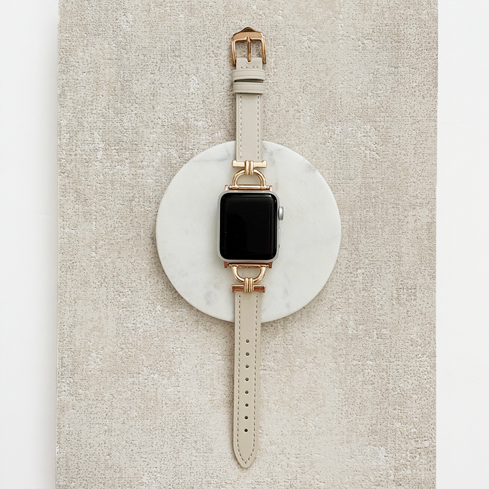 Vegan PU leather Apple Watch strap in oat with gold hardware, displayed on a round marble-textured surface.