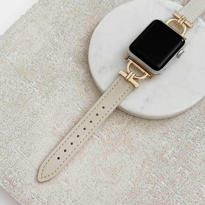 Oat-colored vegan PU leather Apple Watch strap with gold hardware displayed on a round white marble tray.