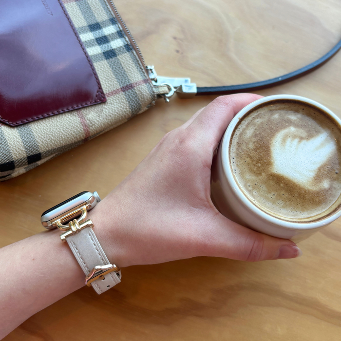 Vegan PU leather Apple Watch strap in oat next to a plaid bag and a hand holding coffee on a wooden table.