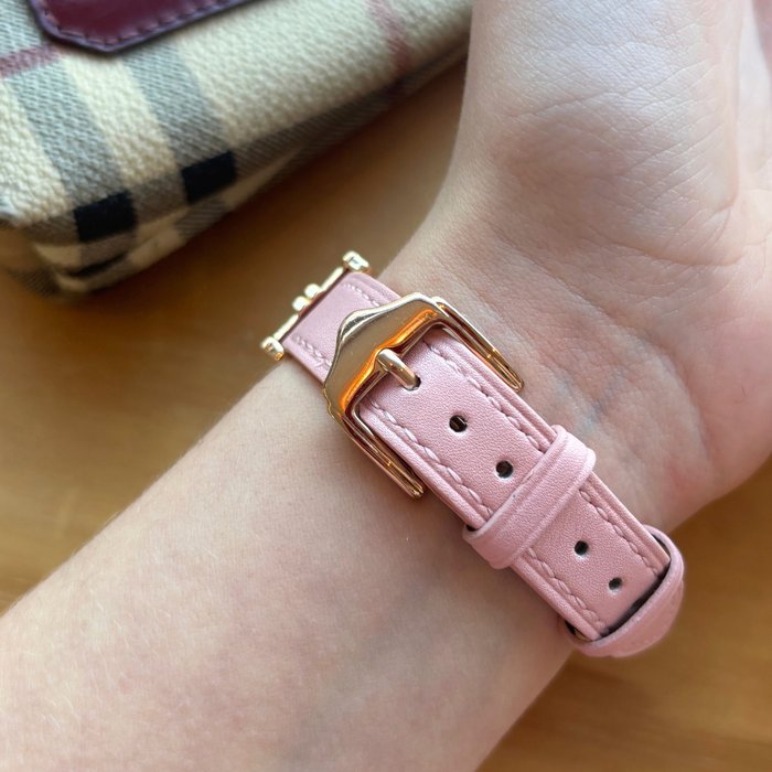 Powder pink vegan leather Apple Watch strap with gold buckle on wrist, next to a plaid fabric item.