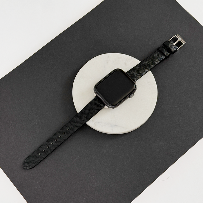 Black vegan textured PU leather Apple Watch strap displayed on a white platform against a black background.