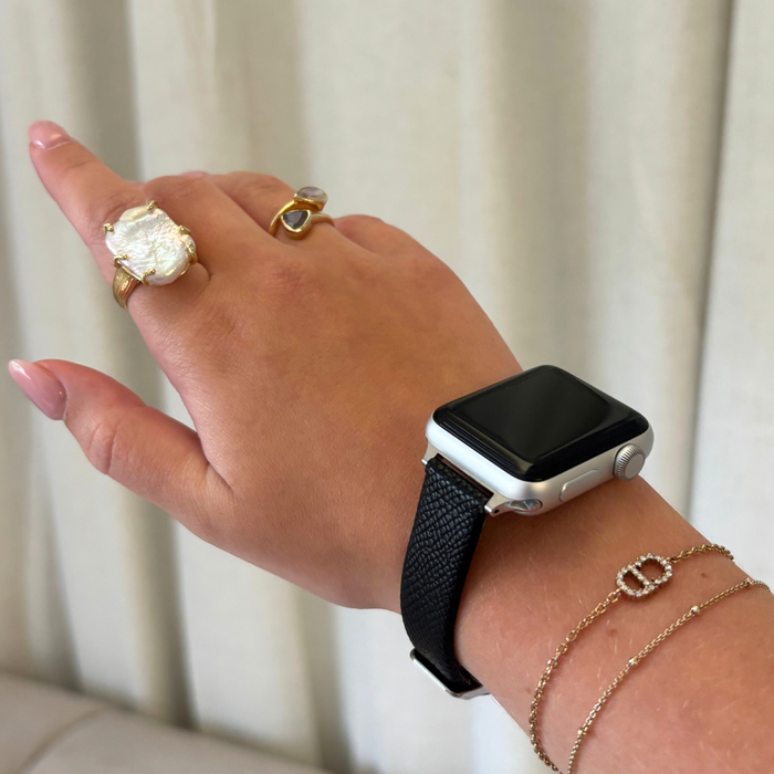 Black vegan leather Apple Watch strap on wrist with gold rings and chain bracelets against a light background.