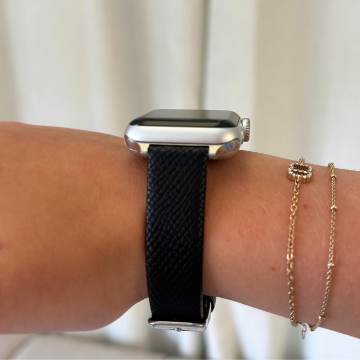 Wrist with a black textured vegan leather Apple Watch strap from Coconut Lane, paired with two gold bracelets.